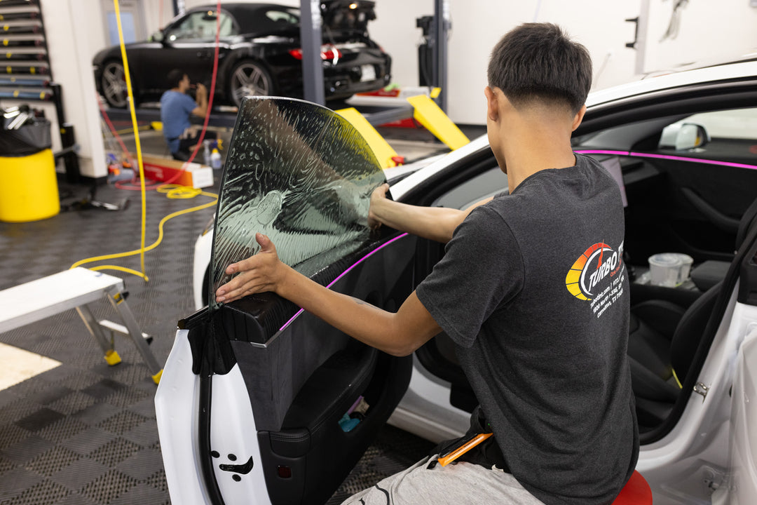 professional installer in a gray t-shirt applies a large, heavily textured clear protective film to a black car door panel inside a bay window tint turbo tint shop.
